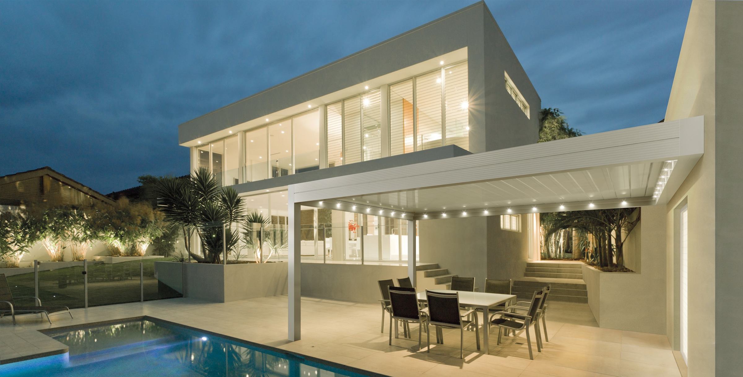Weiße Lamellenpergola mit LED-Licht am Pool einer modernen Villa in der Abenddämmerung